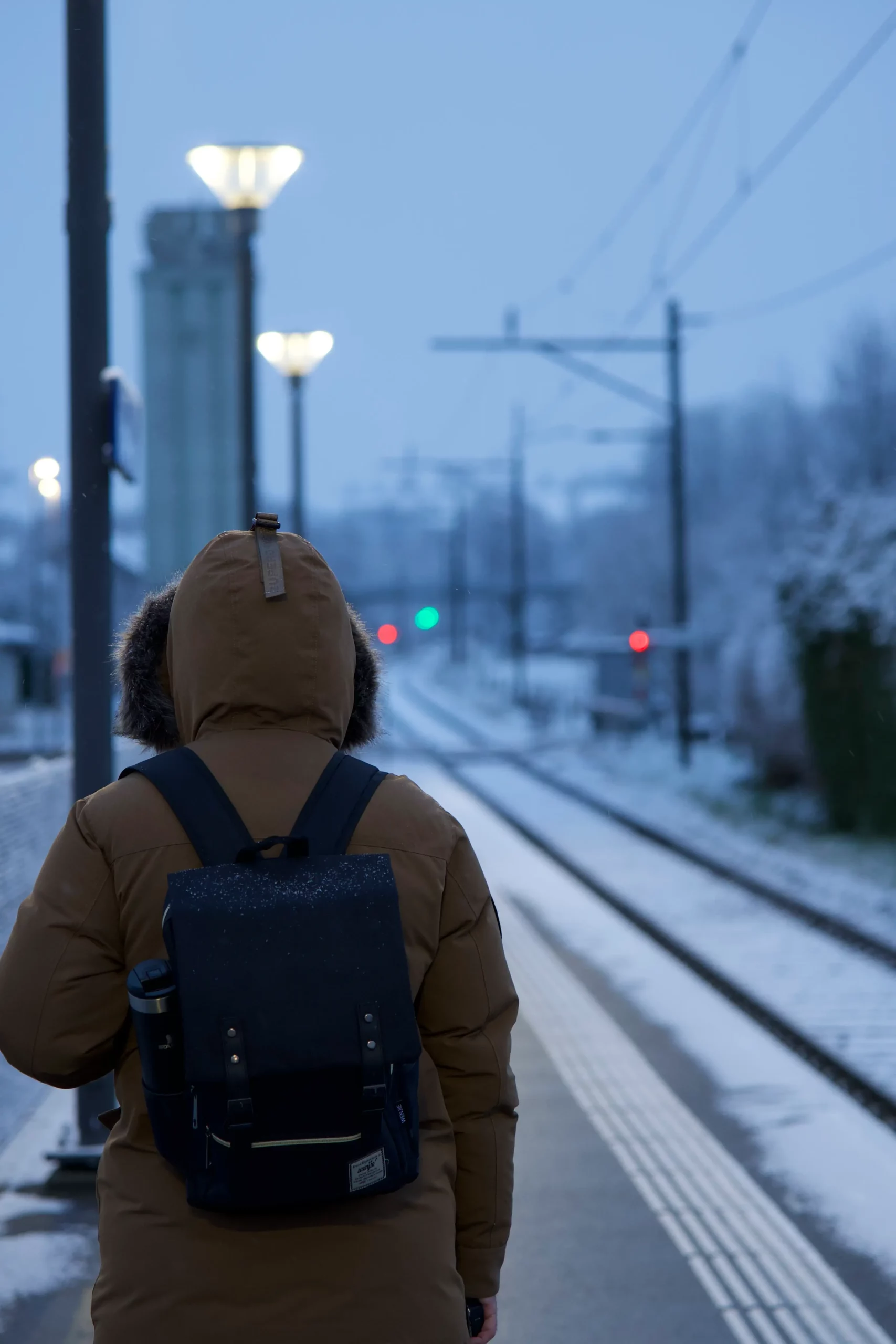 Femme dos parka quai gare train neige lumières crépuscule, objectif grand-angle street urban mélancolique.