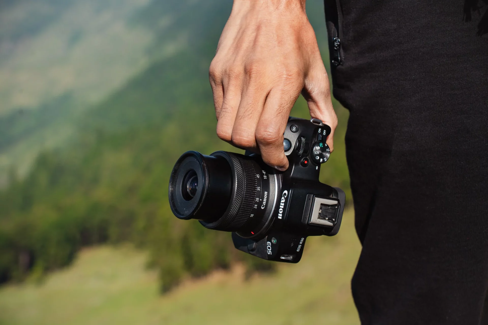 Main tenant un appareil compact Canon EOS R50 devant un paysage de montagne flou.